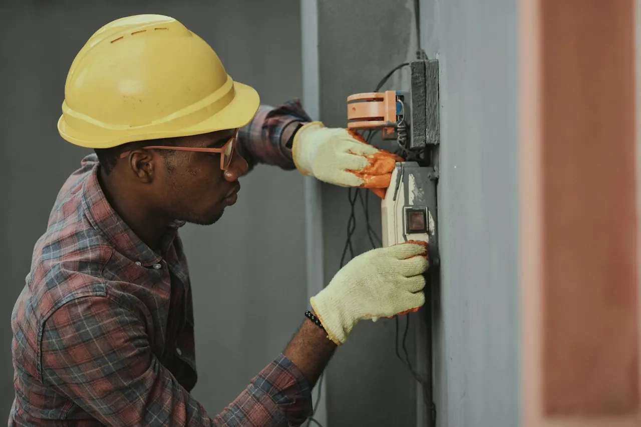 An electrician in a hard hat and gloves working on a wall panel